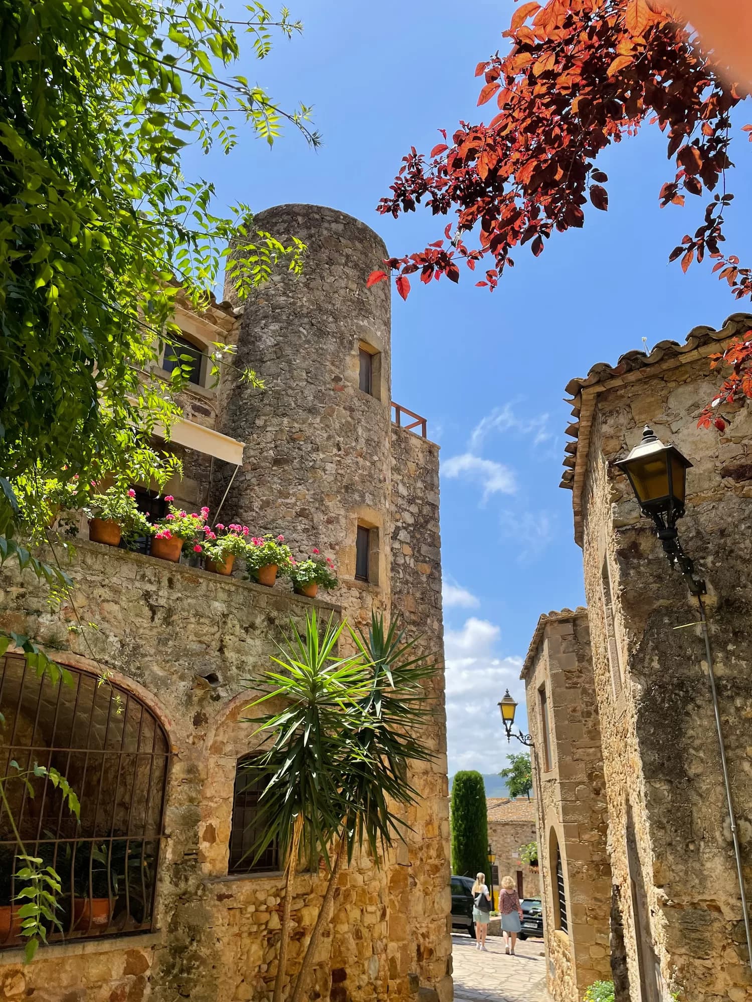 Una calle empedrada de Pals entre casas de piedra, con buganvillas y una torre medieval al fondo